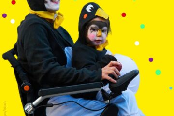 Person im Rollstuhl mit Kind auf dem Schoß. Beide sind als Pinguine verkleidet. Gelber Hintergrund mit bunten Konfetti-Punkten.