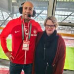 Mann und Frau. Er hat ein Headset auf. Im Hinterrund ein Fußball Stadion.